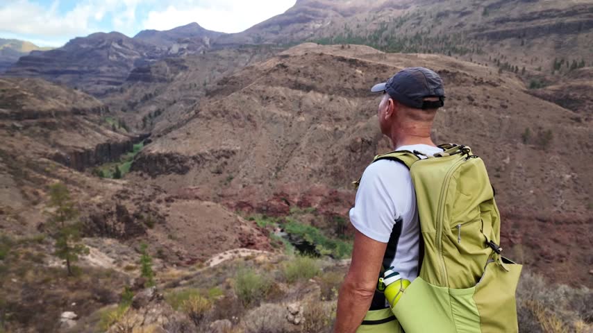 Slow motion footage of a mature adult man with a backpack standing on a cliff edge, peacefully admiring the stunning view of an arid mountain canyon, embracing a moment of freedom and solitude