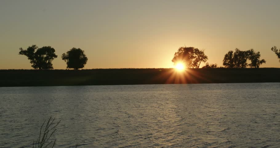 Sun sets behind silhouetted trees on levee overlooking rippling river water   