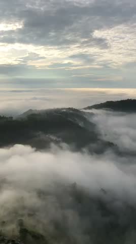 Aerial view of rolling green hills covered by a thick, moving blanket of low clouds and morning mist under an overcast sky.