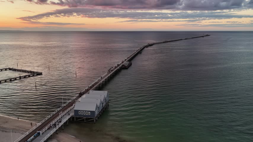 Aerial drone view of Busselton Jetty at sunset, Geographe Bay, Western Australia. Warm light spreads across a glassy bay. A landmark of Western Australia