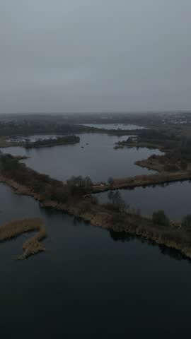 Drone footage glides above interconnected wetlands and calm ponds, revealing reed lined islands, wooded banks, and a distant town beneath gray skies