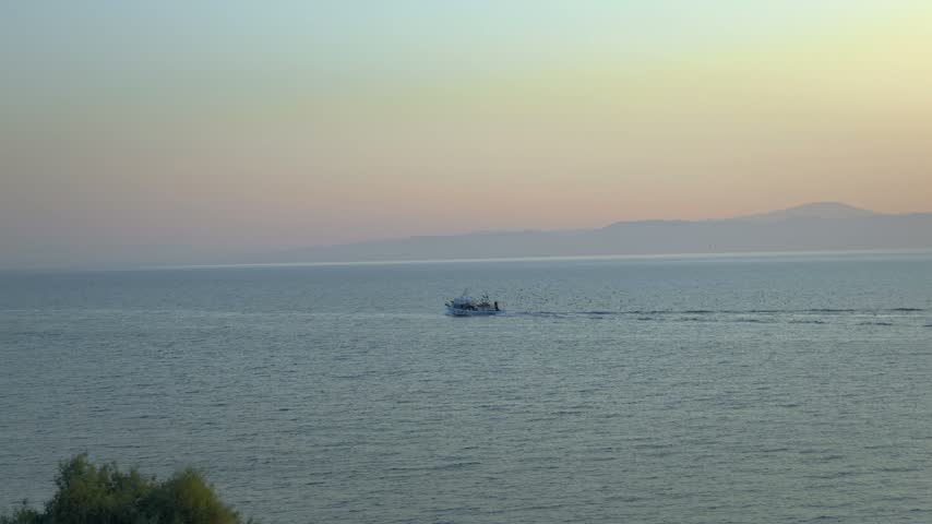 Stunning twilight coastal view of the Aegean Sea, a traditional fishing boat cruising calm waters accompanied by a large flock of seabirds, Mytilene, Lesvos, Greece.