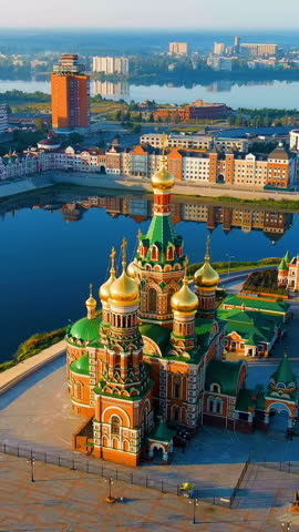 Vertical video. Yoshkar-Ola, Russia. The Cathedral of the Annunciation of the Blessed Virgin Mary stands prominently on the embankment of the Malaya Kokshaga River, its Russian Revival architecture distinguished by five gilded onion domes and richly patterned brickwork. Built between 2009 and 2014 and consecrated in 2016, the cathedral was designed to resemble the Church of the Savior on Spilled Blood in St. Petersburg, making it a striking centerpiece of Republic Square. The aerial perspective captures the cathedral
