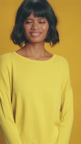 Asian individual with bob haircut clenches fists and smiles broadly, showing intense excitement and triumph against solid yellow backdrop. Enthusiastic energy defines this happy, vibrant moment