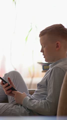 A redhaired boy in blue pajamas is using his phone while lying on the sofa in the evening.