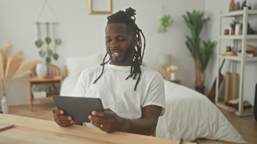 Man holding tablet and tapping screen, laughing with hand to face and smiling at device in tidy bedroom setting; relaxation joy.