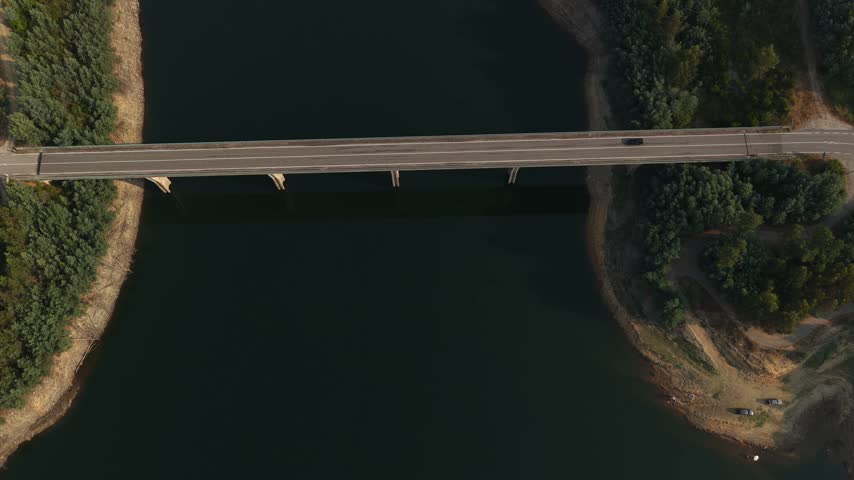 Drone video captures a black car driving across a high bridge spanning a calm river, surrounded by dense greenery and winding road in soft, golden hour lighting.
