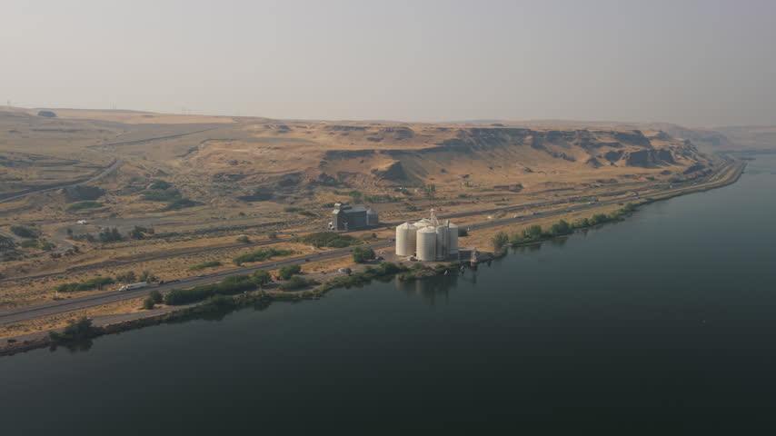 Grain silos and agricultural storage facility beside the Columbia River and Interstate 84 in Oregon, with wildfire smoke creating hazy conditions across the dry landscape.