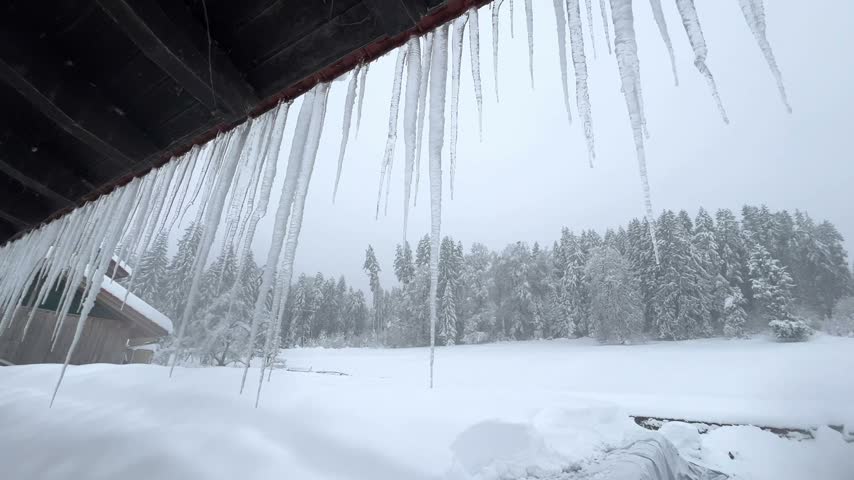 Heavy snowfall blanketing a serene winter landscape, with long icicles hanging from the roof of a cozy wooden cabin in a snowy forest during a cold day in the Bavarian Alps