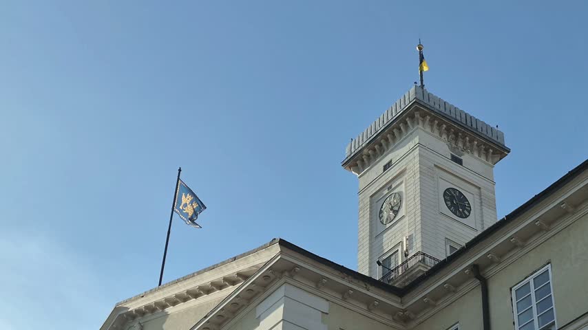 ​Historic clock tower of Lviv Town Hall with the official blue city flag waving under a clear blue sky in Rynok Square, Ukraine.