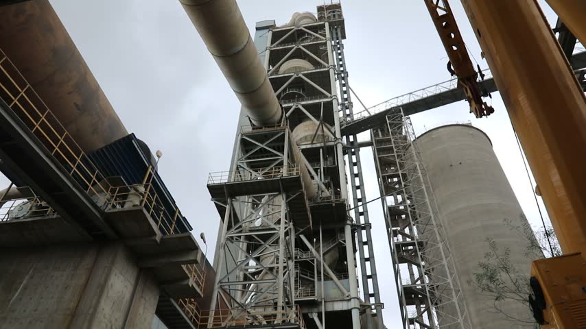 Tilt-down camera shot showing a industrial cement factory under construction, featuring preheater towers, storage silos, and heavy crane machinery.