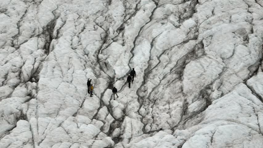 An aerial tracking shot pans right over hikers exploring the rugged Falljökull glacier on Iceland. The vast ice formations and volcanic ash create a stunning, remote landscape.