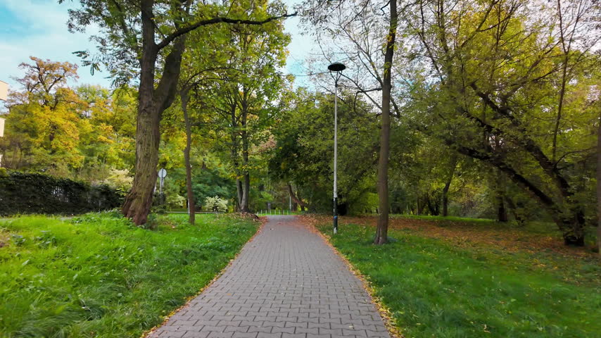 Continuous smooth motion video moving along a paved stone pedestrian walkway in a modern public park surrounded by autumn trees on a sunny day.