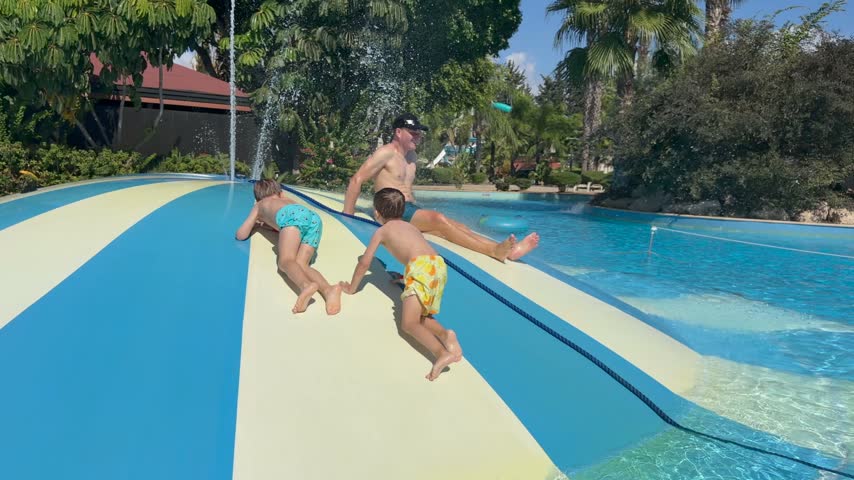 Two boys climb water slide together beside swimming pool, with adult man in water nearby. Children laugh and crawl uphill on wet striped surface beneath palm trees in bright sunshine.