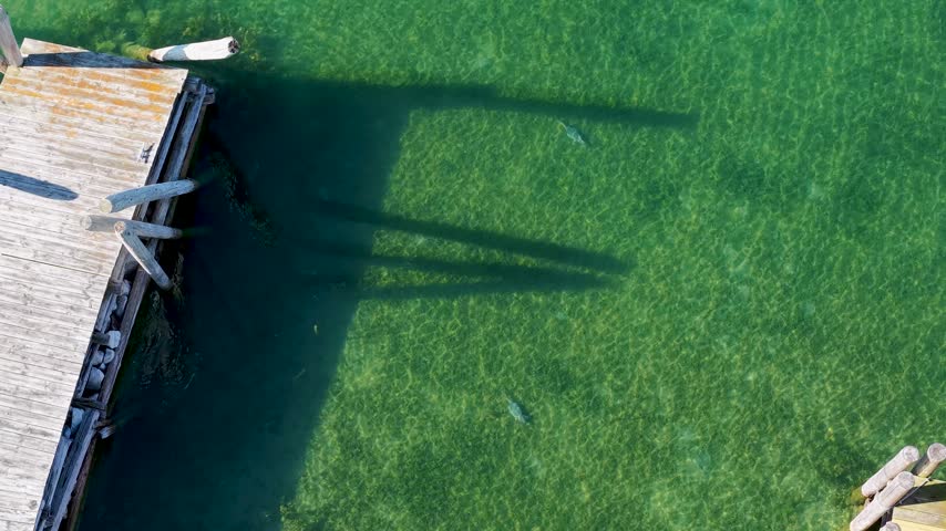 A straight down drone view shows a weathered wooden dock casting long shadows into clear green Lake Huron water at Hessel.