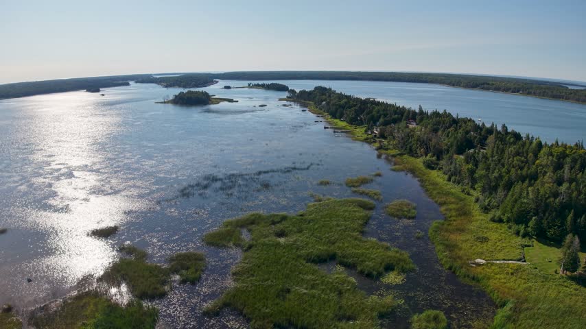 A wide drone view shows reflective shallow wetlands beside a long forested peninsula and small islands near Hessel Michigan.