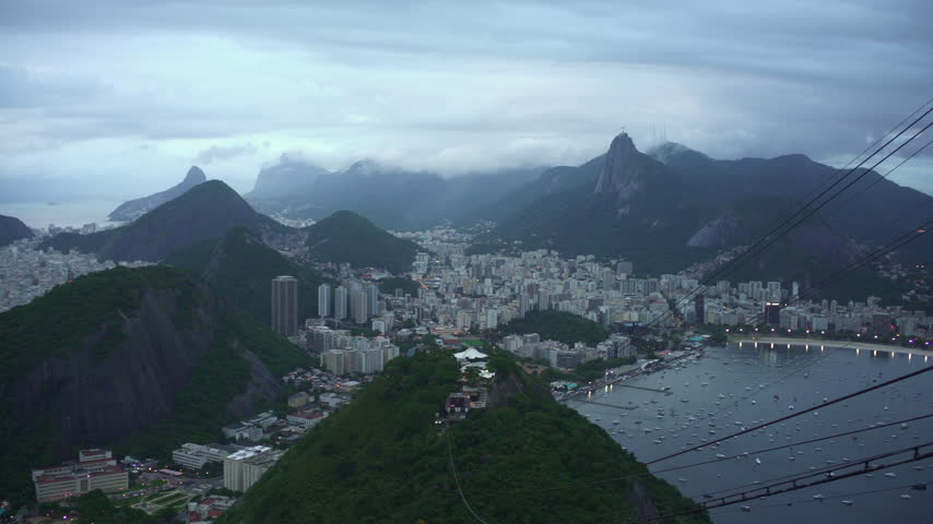 The camera pans from Botafogo Bay across the city and green hills to Copacabana Beach and the ocean under a cloudy sky, seen from Sugarloaf Mountain in Rio de Janeiro, Brazil, Pan