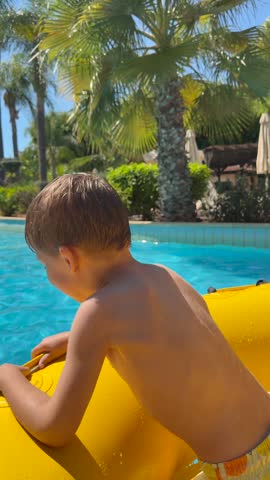 Young boy secures yellow inflatable raft at sunny swimming pool, bending forward to handle its strap. Attentive child steadies float beside clear blue water and tropical greenery, holding it in place.