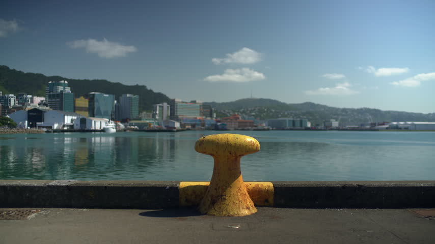 A yellow mooring bollard stands on the quay edge facing Wellington Harbour and the city as two women walk past in the foreground, New Zealand