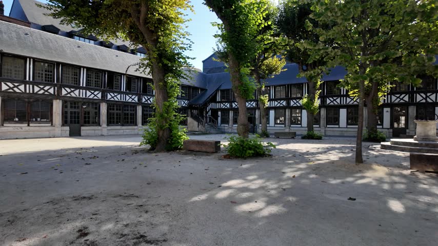 Courtyard of Aître Saint-Maclou in Rouen, France with half-timbered buildings, mature trees, stone paving and shaded outdoor space