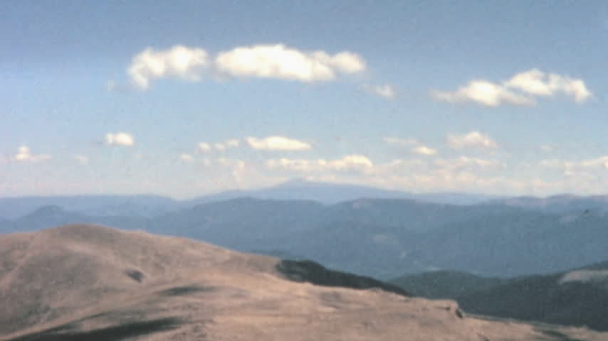 Vast Rocky Mountains landscape looking across barren alpine tundra to layered forested ranges. The view slowly zooms in on a distant prominent hazy peak under a blue sky in Colorado archival footage.