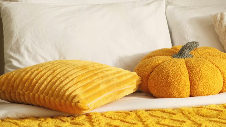 A woman arranges pillows on a bed with white sheets and a textured yellow throw; there are yellow and white plush pillows and an orange pumpkin-shaped, cozy fall bedroom decor in warm morning light