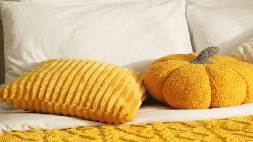 A woman arranges pillows on a bed with white sheets and a textured yellow throw; there are yellow and white plush pillows and an orange pumpkin-shaped, cozy fall bedroom decor in warm morning light