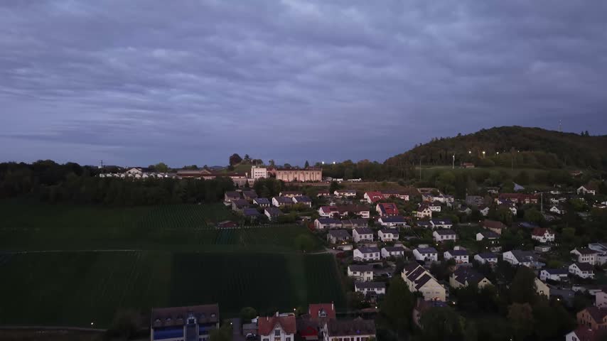 Aerial view of Schaffhausen, Switzerland, at dusk, showing vineyards on green hillsides, residential houses, trees and the Rhine River beneath a cloudy evening sky.