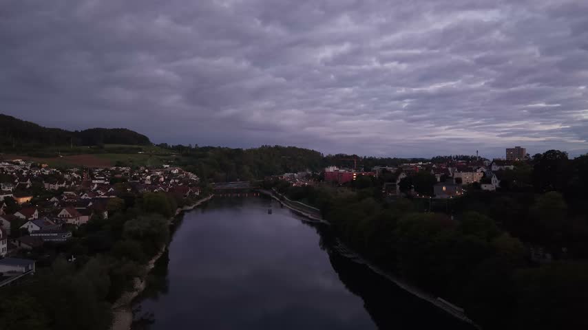 Aerial view of Schaffhausen, Switzerland, at dusk, showing the Rhine River, residential neighborhoods, a bridge and wooded hills beneath a dramatic cloudy evening sky.