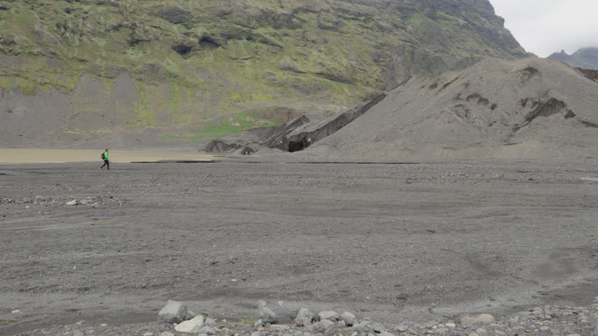 A person in a green jacket hikes across a vast, bleak volcanic plain on Iceland, moving towards a glacier terminus and rugged mountains. This remote landscape evokes adventure and solitude.