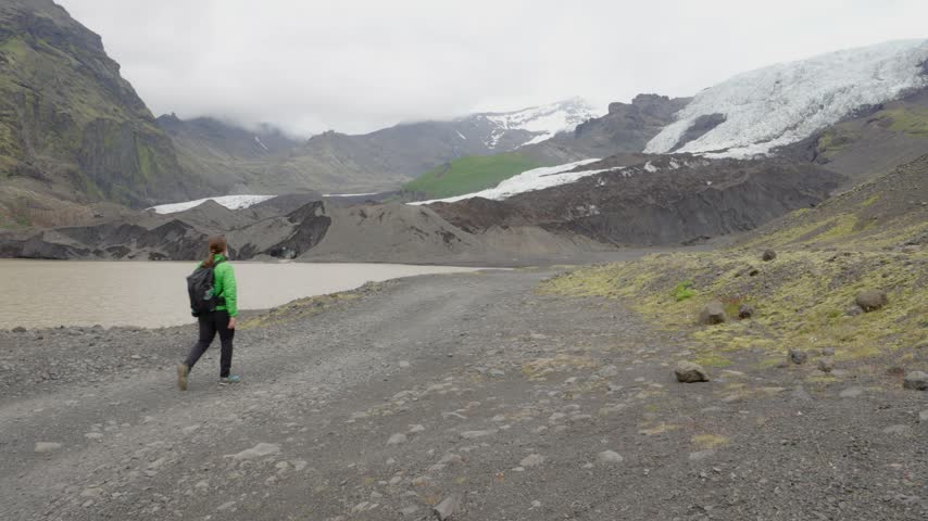 A lone hiker walks on a gravel path alongside a vast glacier lagoon with majestic mountains and an ice cap in the distance on Iceland. Capturing the rugged, remote, and awe-inspiring natural beauty.