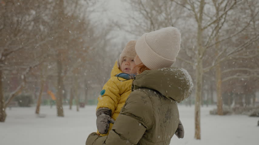 A joyful moment unfolds as a mother and her child experience soft snowfall in a serene park. They gaze up, captivated by the beauty of winter