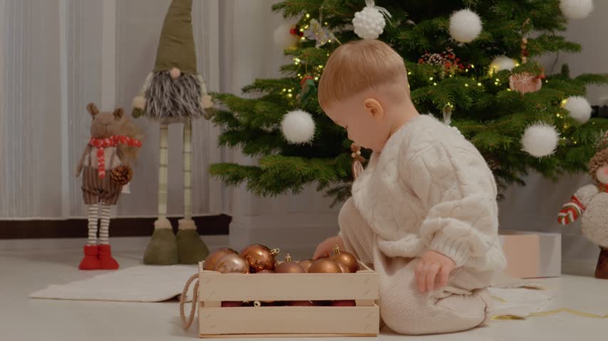 A little boy sits on the floor and takes out ornaments from a box. He smiles while playing with colorful decorations near a Christmas tree in the living room.