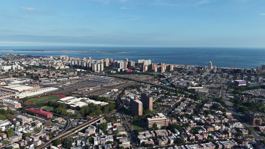 New York City rooftops cluster densely across a coastal neighborhood with rail yards and a school track field near sandy barrier-island beaches along the Atlantic Ocean. Aerial drone wide shot.