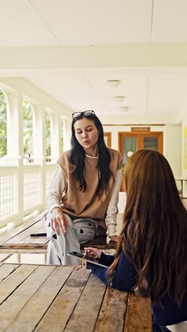 Two women sit at a wooden table in a bright corridor; one looks upset while the other listens, offering support during an emotional conversation.