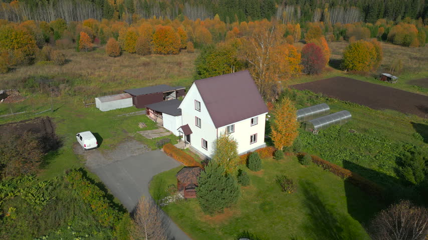 Autumn rural farmhouse with white house, brown roof, garden gazebo, white van, outbuildings, green lawns, cultivated fields, and colorful forest edge under soft daylight from high aerial view