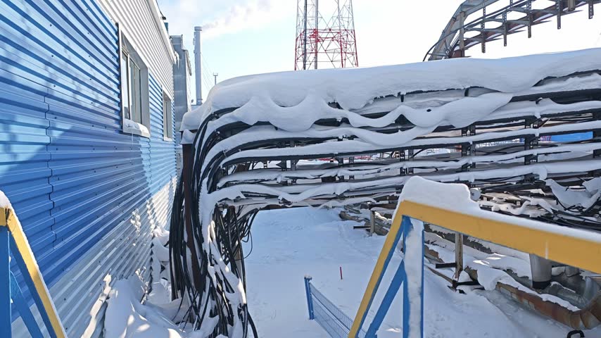 Snowy industrial site with thick cables and pipe runs under winter snow. Utility installation looks quiet and frozen, with fences, rails, and steel supports defining cold facility.