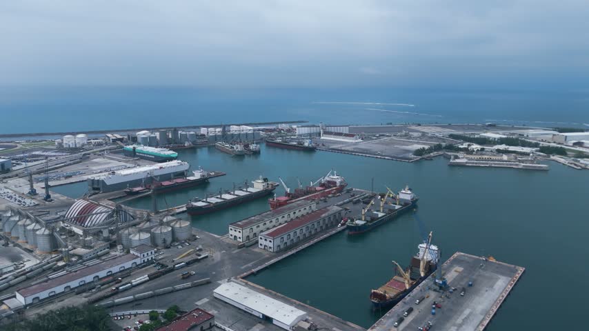 Veracruz, Mexico, a high aerial view looks over the industrial docks of the Port of Veracruz, where cargo ships, bulk carriers and car carriers are moored along long concrete piers. Grain silos, warehouses, storage tanks and railway lines fill the port area, while cranes load and unload goods at the quays. Founded in 1519, Veracruz is the oldest port in Mexico and today moves a large share of the trade between the country, North America and Europe. Beyond the harbor, a breakwater separates the calm water from the open Gulf of Mexico under a gray overcast sky, showcasing the scale of one of the busiest commercial ports in Latin America.