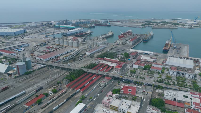 Veracruz, Mexico, an aerial view moves over the port district of Veracruz, where a wide railway yard with freight trains, a road bridge and rows of red roofed warehouses leads toward the docks. Grain silos and storage tanks rise beside the quays, where cargo ships and bulk carriers are moored beneath cranes in the calm harbor. Founded in 1519, Veracruz is the oldest port in Mexico and today moves a large share of the trade between the country, North America and Europe. The industrial port stretches toward the open sea under a soft hazy sky, showcasing the infrastructure that connects rail, road and maritime trade on the Gulf of Mexico.