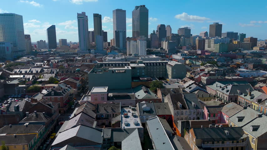 An aerial view of the French Quarter rooftops and downtown New Orleans skyline. A high-angle drone perspective captures the historic pitched rooftops, dormers, and colorful facades of the French Quarter stretching toward the modern skyscrapers of downtown New Orleans. The daytime shot highlights the vivid architectural contrast between the low-rise historic district and the towering Central Business District under a bright blue sky. 