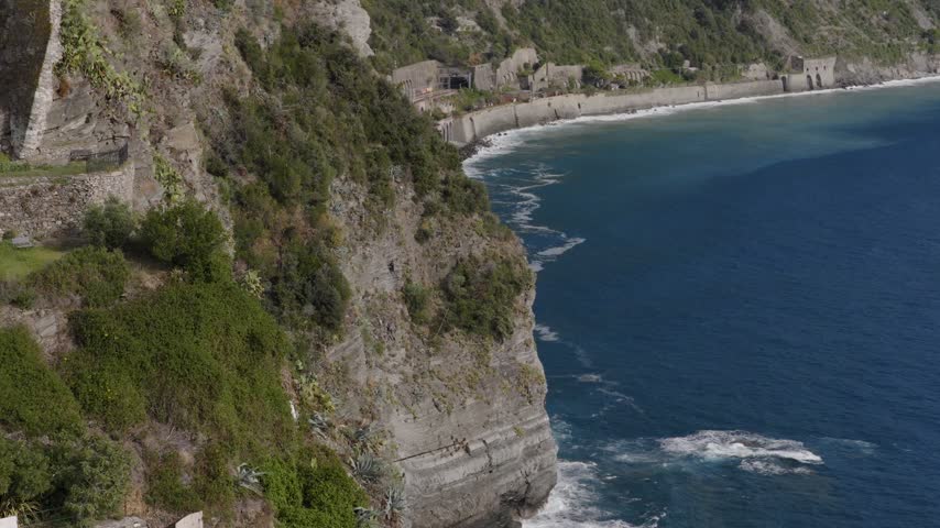 Aerial view of the rugged cliffs of Corniglia, featuring lush Mediterranean vegetation, steep rocky slopes, and the deep blue Ligurian Sea in Corniglia, Liguria, Italy.