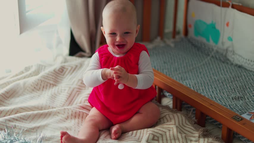 A cheerful infant turns beside a wooden crib in a softly lit bedroom, wearing a bright red outfit and reaching toward the camera from the bed