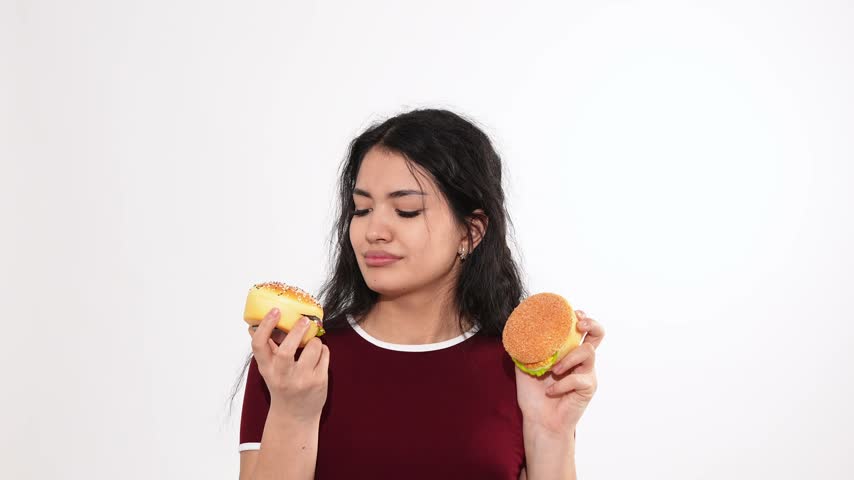 A young woman stands against a white background holding two burgers. She thoughtfully compares them before choosing one, smiling, and taking a bite.