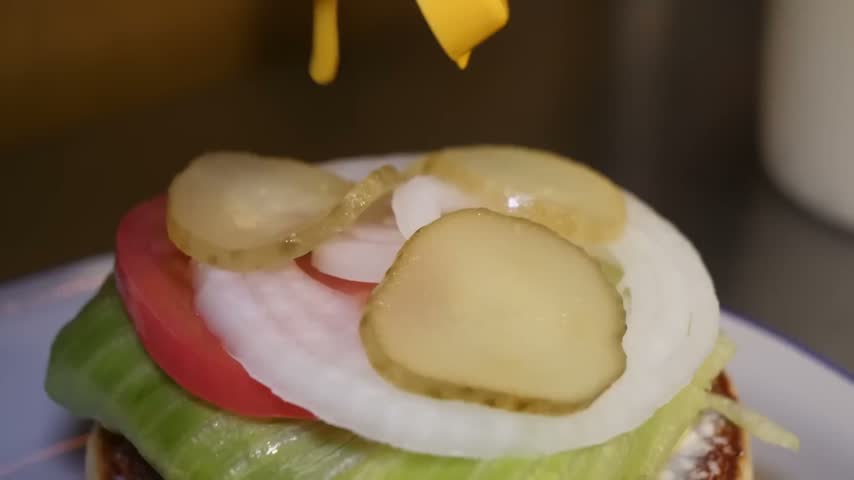 A close-up, slow-motion shot of classic yellow mustard being squeezed from a bottle onto a freshly prepared burger.