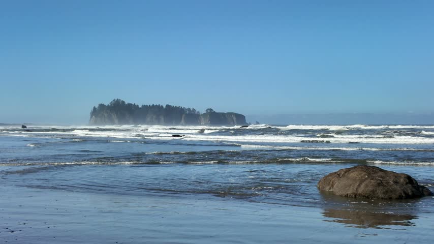 Scenic view of ocean waves on the shoreline with a forested sea stack in the distance at Rialto Beach, Washington