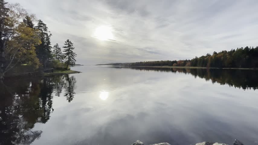 A serene and atmospheric landscape featuring a wide, glassy lake perfectly reflecting a bright sun filtering through an overcast sky. The banks are lined with dark, dense pine and coniferous forests, creating a symmetrical view that evokes a sense of peace, solitude, and untouched wilderness.