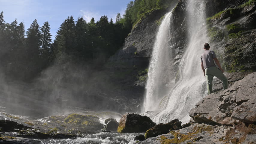A person stands on sloping rocks beside a broad cascade with water spray and forest filling the alpine setting.