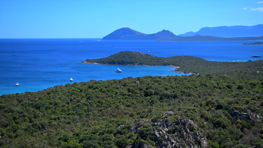 A green peninsula extends into deep blue water with small boats and a hazy mountain profile beyond.