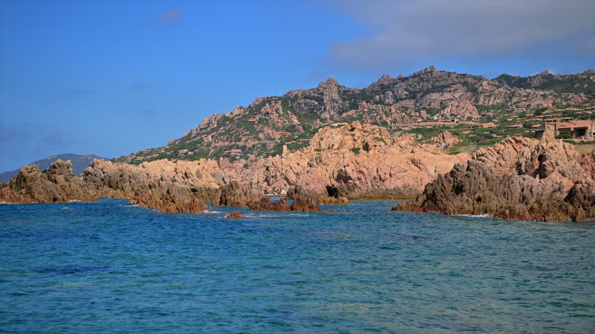 Low coastal rocks rise from the water below a rugged hillside with scattered buildings and green vegetation.