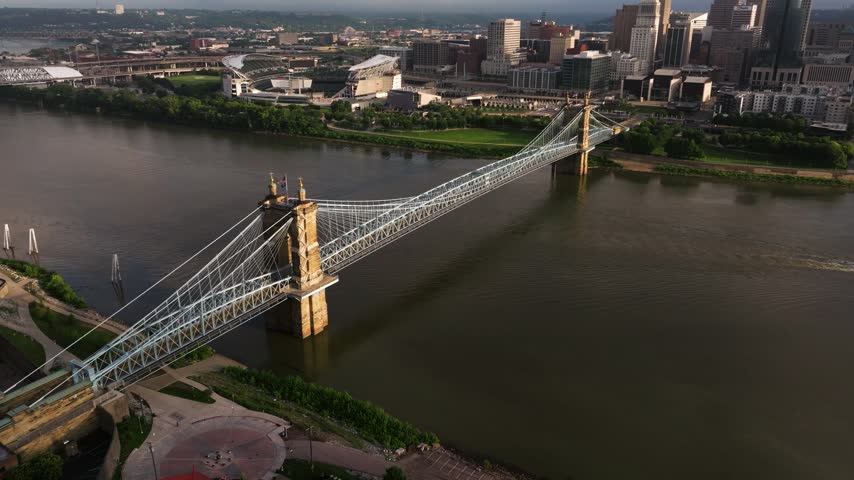 John A. Roebling Suspension Bridge spans the Ohio River toward the downtown Cincinnati skyline, aerial angled dolly at sunset